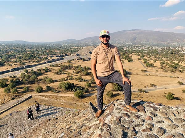 Photo showing Ray standing on a rocky peak overlooking an expansive archaeological site with pyramids and pathways.