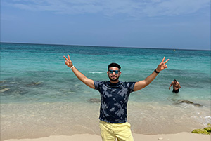 Photo of Ray standing on a sandy beach with the ocean and a clear blue sky in the background