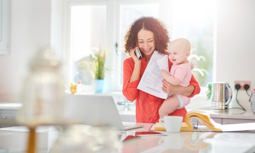 Woman in her kitchen holding a baby and paying a bill by phone